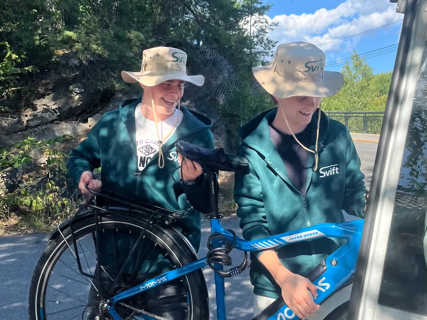 To Svift-ansatte i grønne merkegensere og hatter løfter en blå sykkel inn i varebilen ute på en solrik vei.