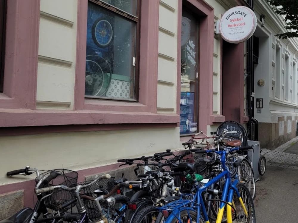 Eiriksgate sykkelverksted på Tøyen i Oslo: en lys bygård med rosa vinduskarmer, verkstedets runde skilt over døra og en rad sykler parkert langs fasaden.